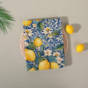 May include: A blue and white floral patterned fabric with yellow lemons and white flowers. The fabric is placed on a woven basket, with three yellow lemons scattered around it. The design is reminiscent of a botanical illustration.