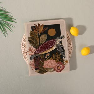 May include: A beige tablet case featuring a sea turtle design. The turtle has a maroon shell and is surrounded by floral and botanical illustrations. A black background with a gold sun shape is behind the turtle. Two yellow lemons are on the right.