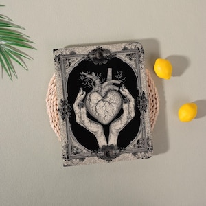 Op de afbeelding: Een zwart-wit notitieboek met een gedetailleerde anatomische hartillustratie, vastgehouden door handen. Het hart staat op een zwarte achtergrond, omlijst door een sierlijke rand. Twee citroenen zijn zichtbaar op de achtergrond.