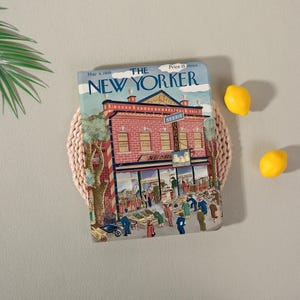May include: A vintage "The New Yorker" magazine cover from May 6, 1939, featuring a detailed illustration of a brick building with a "Seeds" sign. Two yellow lemons rest nearby on a light pink woven mat.