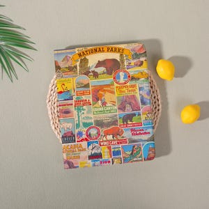 May include: A colorful notebook featuring vintage-style illustrations of various National Parks, including Grand Canyon and Yellowstone. The notebook is placed on a woven placemat, with three yellow lemons nearby. The text "Visit the National Parks" is visible.
