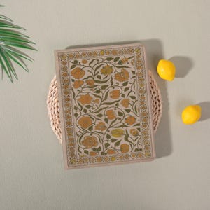 May include: A beige journal featuring a floral pattern in yellow and green. The journal is rectangular with a decorative border. Two yellow lemons are positioned near the journal on a pale green surface.