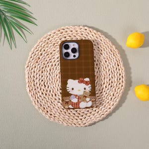 Op de afbeelding: Bruine telefoonhoes met een ruitpatroon en een Hello Kitty-ontwerp. De hoes heeft een witte Hello Kitty met een rode strik en teddyberen. De hoes ligt op een geweven mat met citroenen.