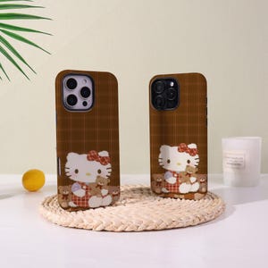 Op de afbeelding: Twee bruine telefoonhoesjes met een ruitpatroon. Elke hoes heeft een stripfiguur met een rode strik en teddyberen. De hoesjes liggen op een geweven mat met een citroen en een kaars op de achtergrond.