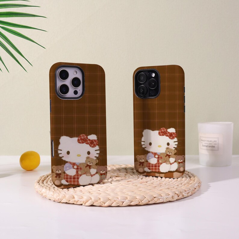 Op de afbeelding: Twee bruine telefoonhoesjes met een ruitpatroon. Elke hoes heeft een stripfiguur met een rode strik en teddyberen. De hoesjes liggen op een geweven mat met een citroen en een kaars op de achtergrond.