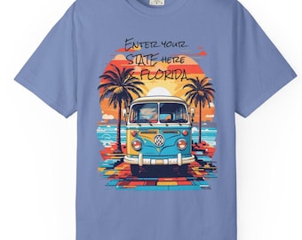 May include: A light blue t-shirt featuring a colorful graphic of a vintage VW bus on a beach at sunset. The graphic includes palm trees, the ocean, and the text "ENTER YOUR STATE HERE FLORIDA".