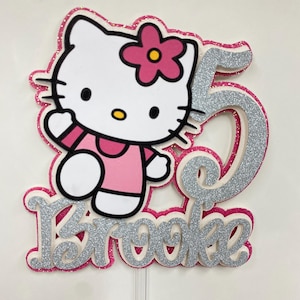 May include: A pink and white Hello Kitty cake topper with a silver glitter number 5 and the name "Brooke". The Hello Kitty figure is wearing a pink dress and has a pink flower on her ear.