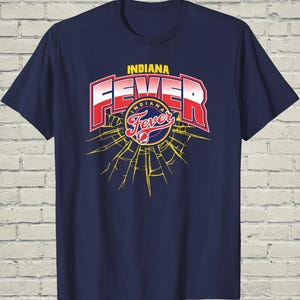 May include: Navy blue t-shirt featuring the text "INDIANA FEVER" in red and white, with a basketball graphic. The design includes a yellow burst pattern. The shirt is a classic crew neck style.