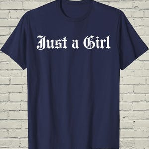 May include: Navy blue t-shirt with the white text "Just a Girl" in a gothic font. The shirt has short sleeves and a crew neck. The background is a white brick wall.