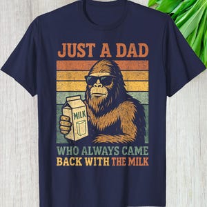 May include: Navy blue t-shirt featuring a retro design with the text "JUST A DAD WHO ALWAYS CAME BACK WITH THE MILK." The graphic shows a Bigfoot character wearing sunglasses and holding a milk carton, set against a striped background.