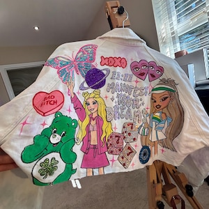 Puede incluir: Chaqueta vaquera blanca con diseños pintados a mano. Presenta una mariposa, un Care Bear y el texto "Manifest Your Dreams". Incluye una figura tipo Barbie y otros elementos gráficos. Una pieza artística y única.