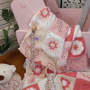 Op de afbeelding: Een roze en witte gehaakte babydeken met bloempatronen, een fopspeen en een houten bijtring. De deken is opgevouwen in een roze houten kinderwagen. Een witte teddybeer is zichtbaar op de voorgrond.
