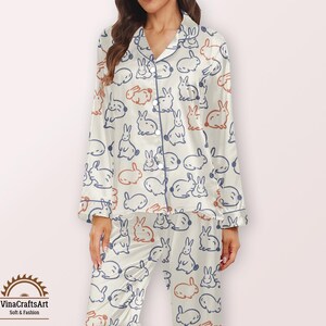 May include: Cream-coloured pyjama set featuring a repeating pattern of cartoon rabbits in blue and orange. The set includes a button-down top with a notched collar and long sleeves, and matching trousers. The pyjamas have blue trim and are made of a soft fabric.