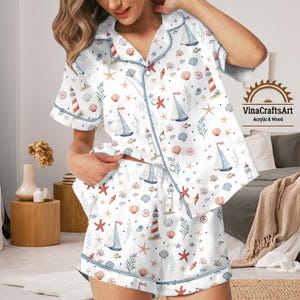 Puede incluir: Conjunto de pijama blanco con temática náutica. El conjunto presenta una camisa de manga corta con botones y pantalones cortos a juego, ambos adornados con veleros, faros, conchas marinas y estrellas de mar en rojo, azul y beige. El ribete es azul claro.