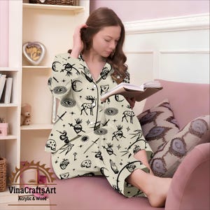 Pode incluir: Conjunto de pijama creme com um padr&atilde;o preto de tema oculto com veados, olhos, luas e estrelas. O conjunto inclui uma camisa com bot&otilde;es e cal&ccedil;as a condizer. O pijama &eacute; usado enquanto se l&ecirc; um livro.