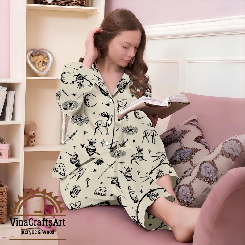 Pode incluir: Conjunto de pijama creme com um padr&atilde;o preto de tema oculto com veados, olhos, luas e estrelas. O conjunto inclui uma camisa com bot&otilde;es e cal&ccedil;as a condizer. O pijama &eacute; usado enquanto se l&ecirc; um livro.