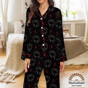 Op de afbeelding: Zwarte pyjamaset met een vleermuis- en maansikkelpatroon. De top met lange mouwen en de broek hebben een herhalend ontwerp in witte en rode accenten. De set heeft rode biezen en witte knopen. Het logo "VinaCraftsArt Soft & Fashion" staat rechtsonder.