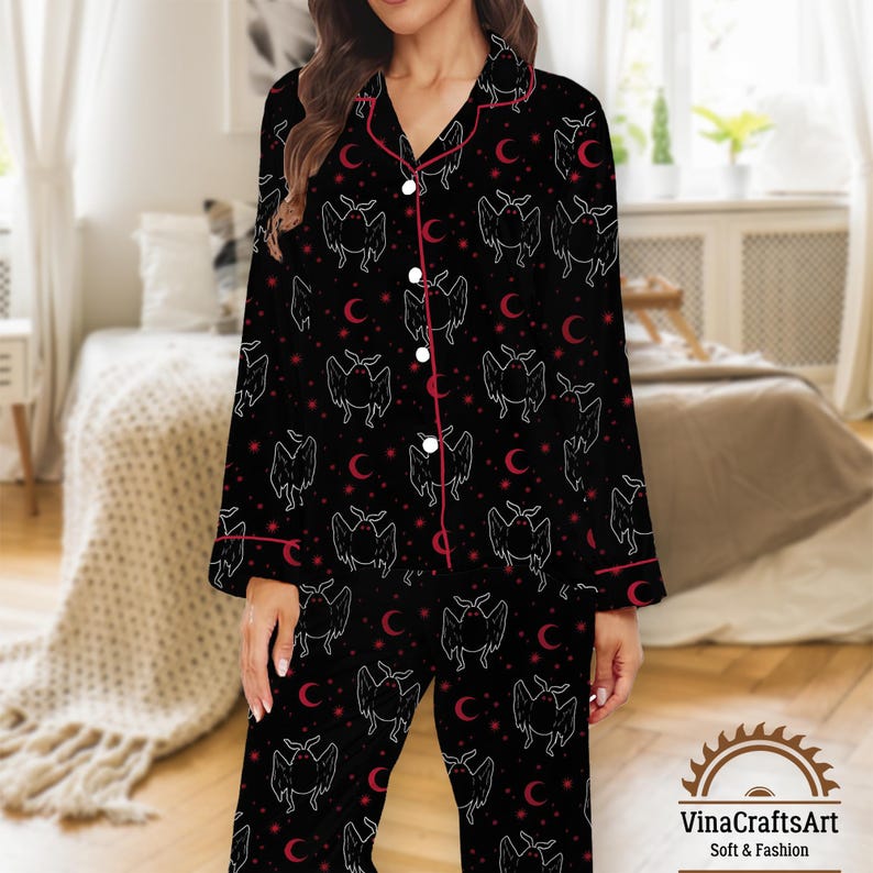 Op de afbeelding: Zwarte pyjamaset met een vleermuis- en maansikkelpatroon. De top met lange mouwen en de broek hebben een herhalend ontwerp in witte en rode accenten. De set heeft rode biezen en witte knopen. Het logo "VinaCraftsArt Soft & Fashion" staat rechtsonder.