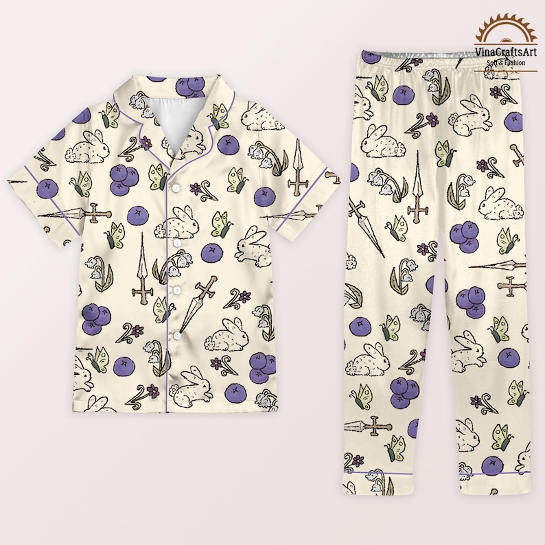 K&ouml;nnte beinhalten: Cremefarbenes Pyjama-Set mit einem sich wiederholenden Muster aus wei&szlig;en Kaninchen, Schmetterlingen, Schwertern und lila Beeren. Das kurz&auml;rmelige Oberteil und die Hose haben einen lila Besatz. Der Pyjama ist aus einem weichen, leichten Stoff.