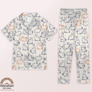 May include: Cream-coloured pyjama set with a repeating pattern of blue and orange bunnies. The set includes a short-sleeved button-down shirt and matching trousers. The pyjamas have a relaxed fit and are made of a soft fabric.
