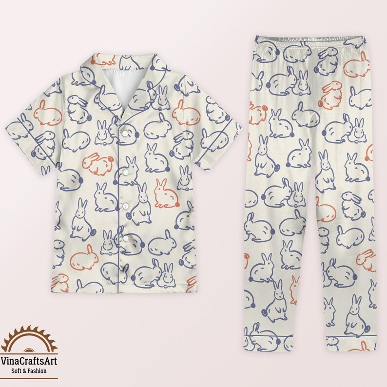 May include: Cream-coloured pyjama set with a repeating pattern of blue and orange bunnies. The set includes a short-sleeved button-down shirt and matching trousers. The pyjamas have a relaxed fit and are made of a soft fabric.
