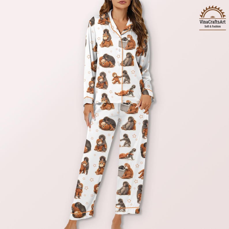 May include: White pyjama set featuring a repeating pattern of brown orangutans and small stars. The long-sleeved top and trousers have orange trim. The pyjamas are made of a soft fabric. The VinaCraftsArt logo is in the upper right corner.