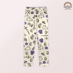 K&ouml;nnte beinhalten: Cremefarbene Schlafanzughose mit einem sich wiederholenden Muster aus wei&szlig;en Kaninchen, lila Blaubeeren, Schmetterlingen und stilisierten Blumen. Die Hose zeigt auch Illustrationen von Schwertern und Kreuzen. Die Schlafanzughose hat eine lila Bord&uuml;re.