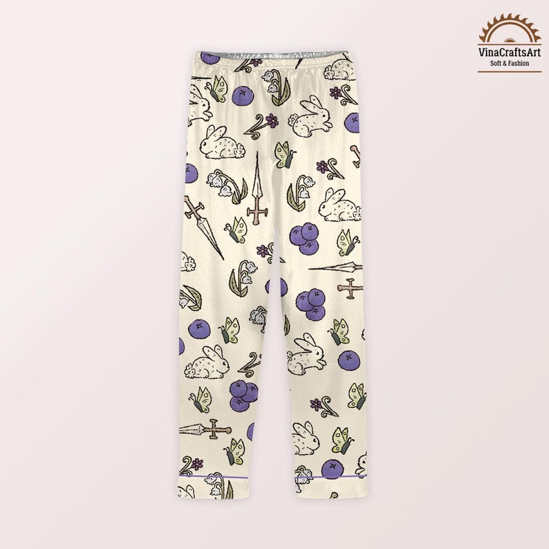 K&ouml;nnte beinhalten: Cremefarbene Schlafanzughose mit einem sich wiederholenden Muster aus wei&szlig;en Kaninchen, lila Blaubeeren, Schmetterlingen und stilisierten Blumen. Die Hose zeigt auch Illustrationen von Schwertern und Kreuzen. Die Schlafanzughose hat eine lila Bord&uuml;re.