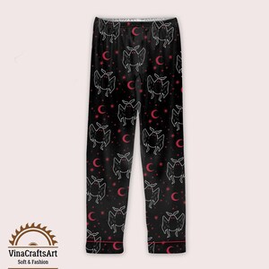 Op de afbeelding: Zwarte pyjamabroek met een herhalend patroon van witte vleermuisachtige wezens, rode halve manen en sterren. De broek heeft rode biezen en de tekst "VinaCraftsArt Soft & Fashion" aan de onderkant.