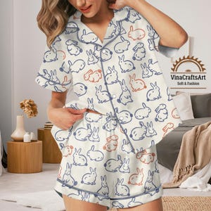 May include: Cream-coloured pyjama set featuring a pattern of blue and orange bunny illustrations. The short-sleeved top and shorts have blue piping. The text "VinaCraftsArt Soft & Fashion" is visible.