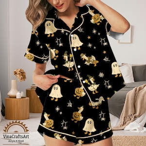 Könnte beinhalten: Schwarzes Pyjama-Set mit Halloween-Motiv und weißen Zierleisten. Das Set besteht aus einem kurzärmeligen Hemd mit Knöpfen und Shorts, beide mit einem sich wiederholenden Muster aus Geistern, Enten, Kürbissen und Spinnweben. Der Markenname "VinaCraftsArt" ist sichtbar.