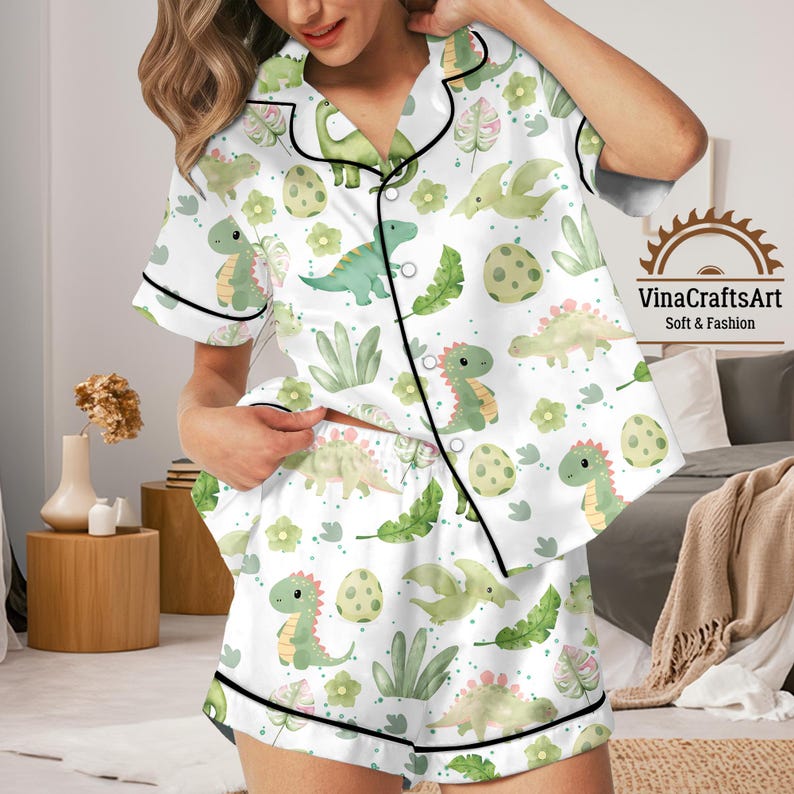 May include: White dinosaur-themed pajama set. The short-sleeved top and shorts are patterned with green dinosaurs, eggs, and leaves. Black trim edges the set. The text "VinaCraftsArt Soft & Fashion" is visible on the right side.