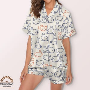 May include: Cream-coloured pyjama set featuring a repeating pattern of blue and orange bunny illustrations. The short-sleeved top and shorts are made of a soft, silky material. The set includes the text "VinaCraftsArt Soft & Fashion".