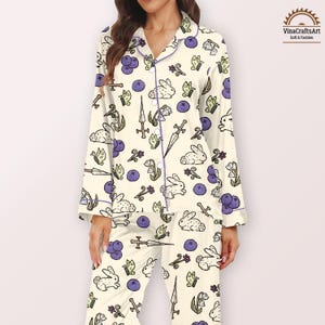 K&ouml;nnte beinhalten: Cremefarbenes Pyjama-Set mit einem skurrilen Muster aus Hasen, Schmetterlingen, Schwertern und Blaubeeren. Das Set hat lila Bes&auml;tze und Kn&ouml;pfe. Das lang&auml;rmlige Oberteil und die Hose sind aus einem weichen Stoff. Das Logo "VinaCraftsArt Soft & Fashion" befindet sich oben rechts.