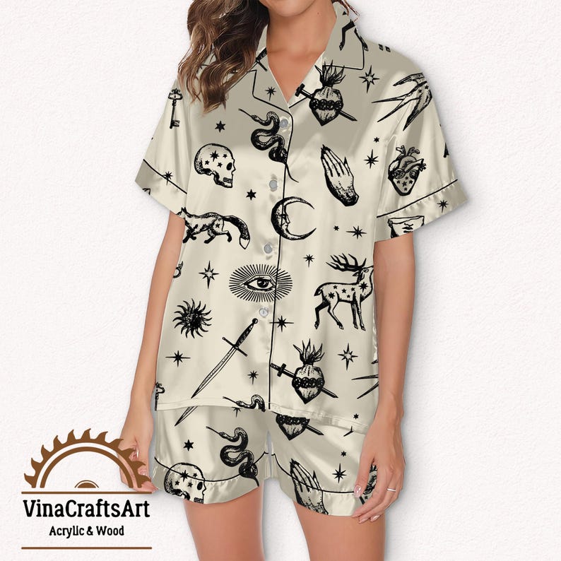 Pode incluir: Conjunto de pijama creme acetinado com um padr&atilde;o oculto preto. O conjunto inclui uma camisa com bot&otilde;es e cal&ccedil;&otilde;es, ambos adornados com s&iacute;mbolos como caveiras, cobras e punhais. O tecido tem um brilho acetinado.