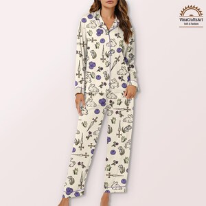 K&ouml;nnte beinhalten: Cremefarbenes Pyjama-Set mit einem sich wiederholenden Muster aus wei&szlig;en Kaninchen, Schwertern, lila Beeren und Blumen. Das lang&auml;rmlige Oberteil und die Hose sind mit lila Paspeln versehen. Der Pyjama ist weich und bequem.