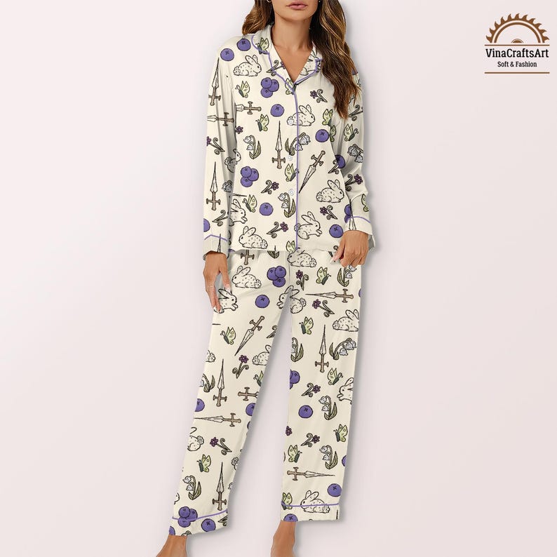 K&ouml;nnte beinhalten: Cremefarbenes Pyjama-Set mit einem sich wiederholenden Muster aus wei&szlig;en Kaninchen, Schwertern, lila Beeren und Blumen. Das lang&auml;rmlige Oberteil und die Hose sind mit lila Paspeln versehen. Der Pyjama ist weich und bequem.