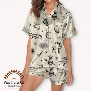 Pode incluir: Conjunto de pijama creme com estampa preta de tema celestial e oculto. O conjunto inclui uma camisa de manga curta com bot&otilde;es e cal&ccedil;&otilde;es a condizer. A estampa apresenta luas, estrelas, espadas e outros s&iacute;mbolos m&iacute;sticos. A marca "VinaCraftsArt" &eacute; vis&iacute;vel.