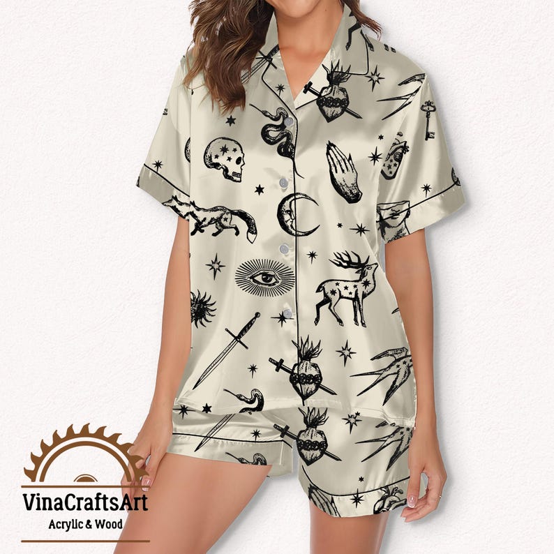 Pode incluir: Conjunto de pijama creme com estampa preta de tema celestial e oculto. O conjunto inclui uma camisa de manga curta com bot&otilde;es e cal&ccedil;&otilde;es a condizer. A estampa apresenta luas, estrelas, espadas e outros s&iacute;mbolos m&iacute;sticos. A marca "VinaCraftsArt" &eacute; vis&iacute;vel.