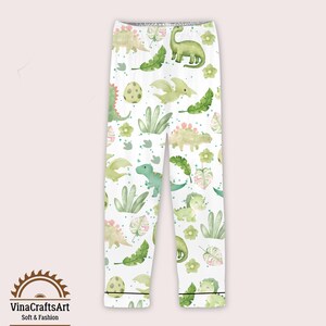 May include: White pajama bottoms featuring a repeating pattern of green and yellow dinosaurs, eggs, and foliage. The trousers have a black trim at the ankles. The text "VinaCraftsArt Soft & Fashion" is at the bottom.
