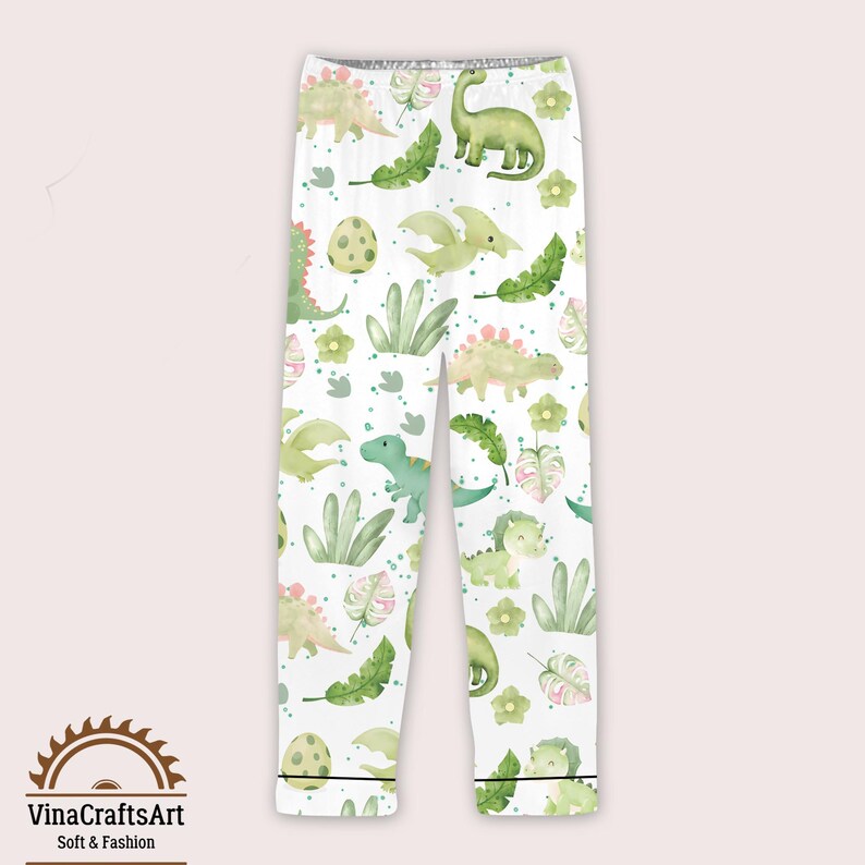 May include: White pajama bottoms featuring a repeating pattern of green and yellow dinosaurs, eggs, and foliage. The trousers have a black trim at the ankles. The text "VinaCraftsArt Soft & Fashion" is at the bottom.