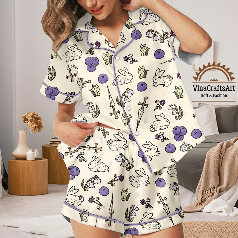 K&ouml;nnte beinhalten: Cremefarbenes Pyjama-Set mit einem skurrilen Muster aus wei&szlig;en Kaninchen, lila Blaubeeren, Schmetterlingen und Schwertern. Das Set umfasst ein kurz&auml;rmeliges Hemd mit Kn&ouml;pfen und passende Shorts, die mit lila Paspeln versehen sind. Der Text "VinaCraftsArt Soft & Fashion" ist sichtbar.