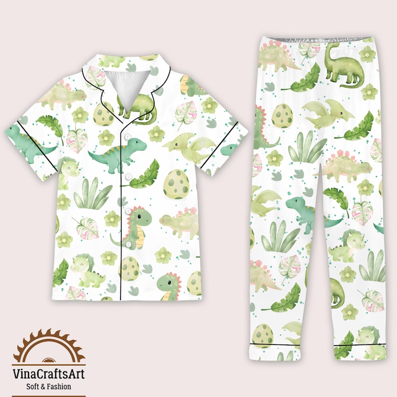 May include: White pajama set with a dinosaur pattern. The short-sleeved top and trousers feature green dinosaurs, eggs, and foliage. The set has black trim and is made of soft material.