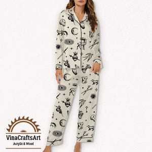 Pode incluir: Conjunto de pijama creme com um padr&atilde;o preto com tema oculto. A parte superior de manga comprida e as cal&ccedil;as apresentam v&aacute;rios s&iacute;mbolos como luas, olhos, cora&ccedil;&otilde;es e tesouras. O conjunto tem acabamentos pretos e uma parte superior abotoada.