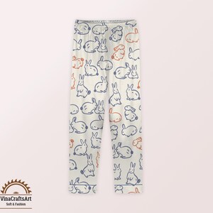 May include: Cream-coloured leggings with a repeating pattern of blue and orange bunny illustrations. The leggings feature a soft and fashionable design, perfect for casual wear. The logo "VinaCraftsArt Soft & Fashion" is visible in the bottom left corner.