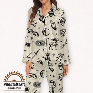 Pode incluir: Conjunto de pijama creme com um padr&atilde;o celestial preto. A parte superior de manga comprida e as cal&ccedil;as apresentam ilustra&ccedil;&otilde;es de luas, estrelas, veados e outros s&iacute;mbolos m&iacute;sticos. O conjunto tem acabamentos e bot&otilde;es pretos, e a marca "VinaCraftsArt" &eacute; vis&iacute;vel.
