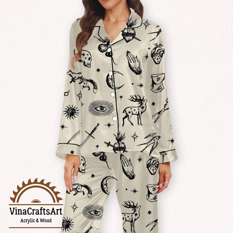 Pode incluir: Conjunto de pijama creme com um padr&atilde;o celestial preto. A parte superior de manga comprida e as cal&ccedil;as apresentam ilustra&ccedil;&otilde;es de luas, estrelas, veados e outros s&iacute;mbolos m&iacute;sticos. O conjunto tem acabamentos e bot&otilde;es pretos, e a marca "VinaCraftsArt" &eacute; vis&iacute;vel.