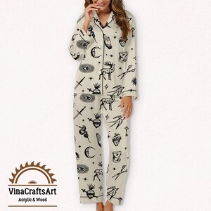 Pode incluir: Conjunto de pijama creme com ilustra&ccedil;&otilde;es pretas de luas, olhos, veados e outros s&iacute;mbolos m&iacute;sticos. A parte superior de manga comprida tem uma gola entalhada e fecho de bot&otilde;es. As cal&ccedil;as a condizer t&ecirc;m cintura el&aacute;stica. O log&oacute;tipo "VinaCraftsArt Acrylic & Wood" est&aacute; na parte inferior.