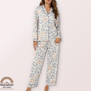May include: Cream-coloured pyjama set featuring a pattern of blue and orange rabbits. The long-sleeved top has a collared neckline and button closure. The pyjama trousers have a matching rabbit print. The brand logo is in the bottom left corner.