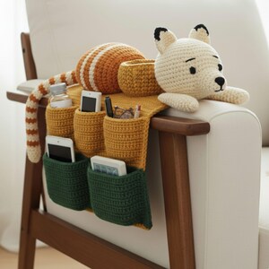 May include: A handmade, crocheted armrest organizer in mustard yellow and green, featuring pockets for storage. The organizer is adorned with a crocheted bear head and a striped tail. It is placed on a white armchair with a wooden frame.